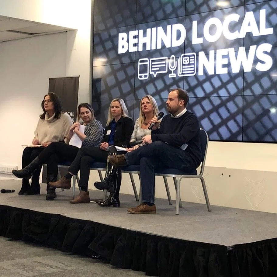 Jono speaking with a microphone on a panel in front of a Behind Local News screen
