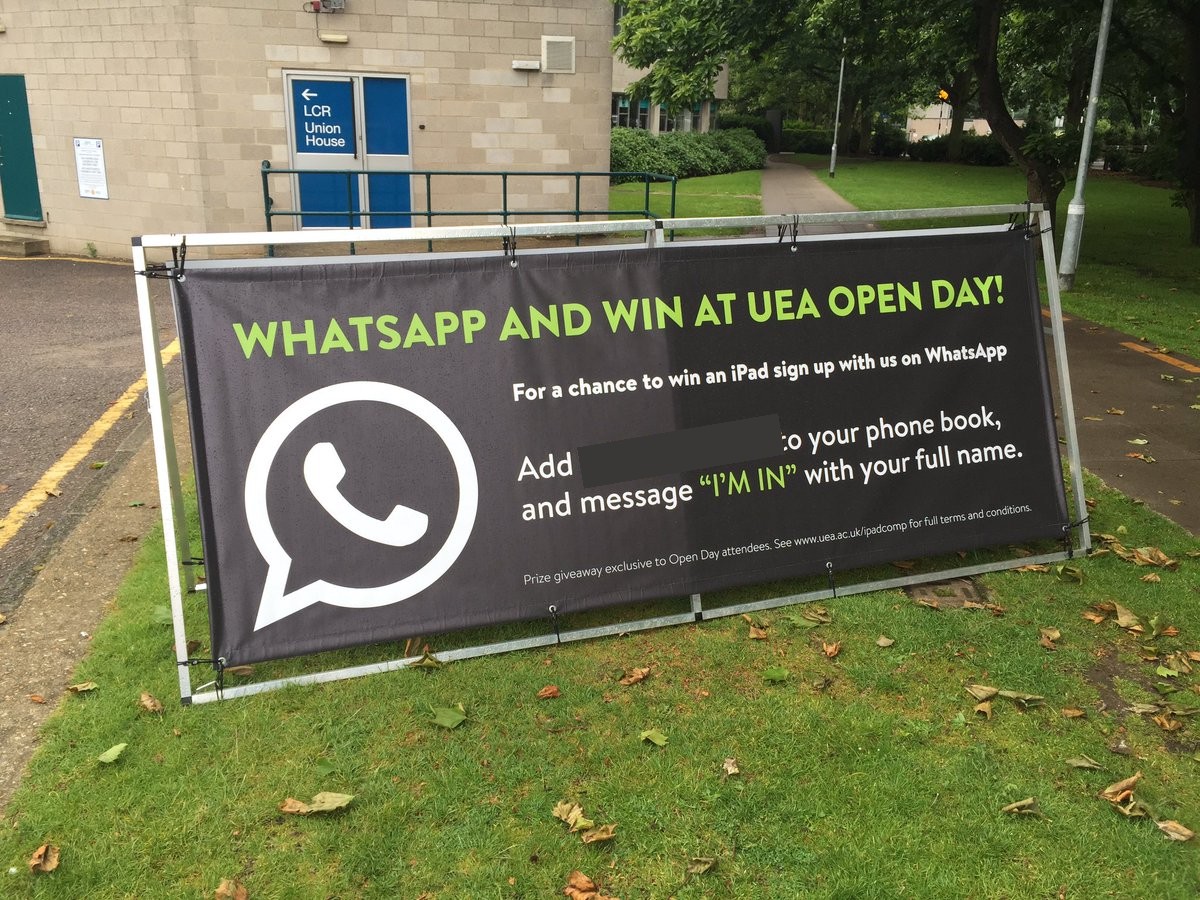 Original University of East Anglia Open Day promotional banner advertising a WhatsApp sign-up competition beside a campus walkway. The historical contact number has been obscured.